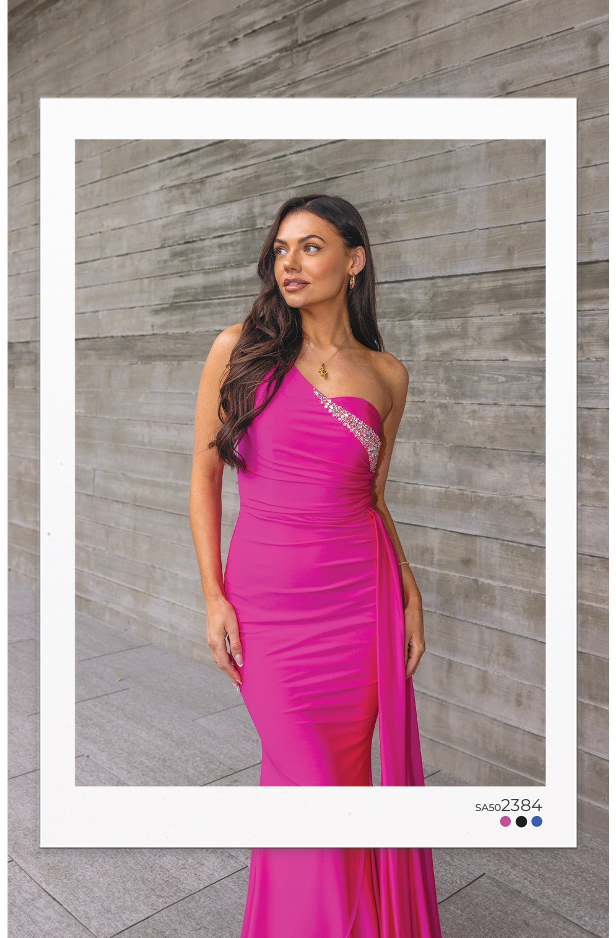 Model in a hot pink one-shoulder fitted evening dress featuring rhinestone neckline detail and flowing side sash against concrete wall.